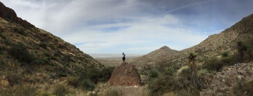A person stands on a rock in a vast desert landscape, surrounded by mountains and a cloudy sky.
