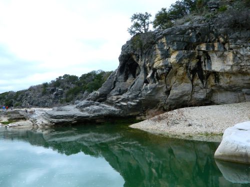 A rocky shoreline with a calm green river reflecting the overcast sky and surrounding trees.