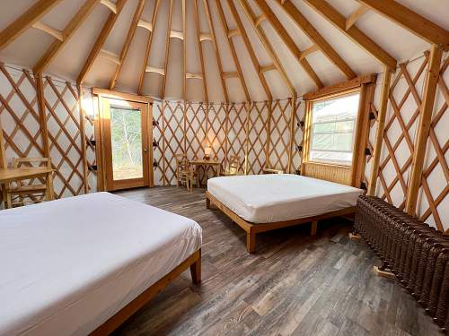 Interior of a cozy yurt with two beds, wooden furniture, and large windows letting in natural light.