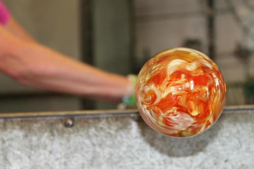 A close-up of a colorful glass orb being shaped, with a hand visible in the background.