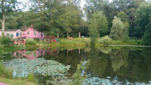 A serene pond reflects a pink house and lush greenery, surrounded by trees and a peaceful landscape.