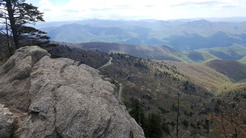 A rocky outcrop overlooks a lush, mountainous landscape with winding roads and distant peaks under a blue sky.