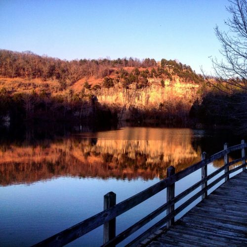 A serene lake reflects a hillside at sunset, with a wooden railing in the foreground and trees lining the shore.