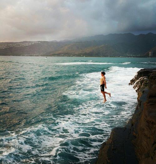 A person stands on rocky shore, facing the ocean with waves crashing, under a cloudy sky.