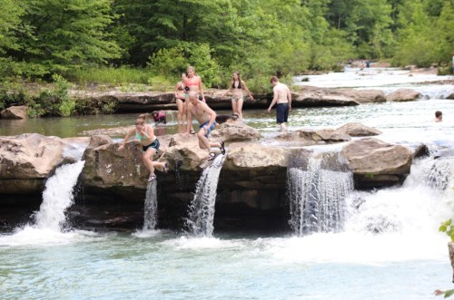 A group of people enjoying a natural swimming area with waterfalls and rocks in a lush green setting.