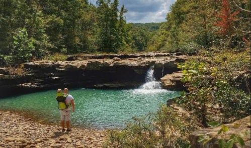 A person stands by a serene, turquoise pool with a small waterfall, carrying a child in a backpack carrier.