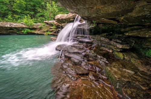 A serene waterfall cascades over rocky ledges into a tranquil turquoise pool, surrounded by lush greenery.