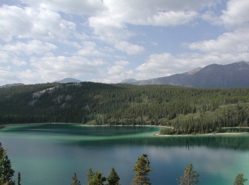 A serene lake surrounded by lush green forests and mountains under a partly cloudy sky.