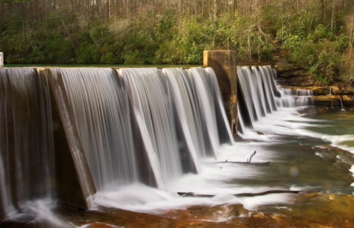 A serene waterfall cascading over a dam, surrounded by lush greenery and a calm river below.