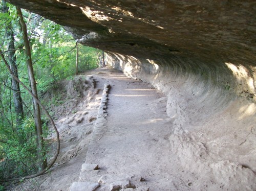 A winding dirt path beneath a rocky overhang, surrounded by lush green trees and natural rock formations.