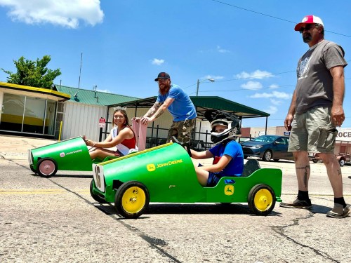 Two children in green toy cars race down a street, while two adults watch and cheer in a sunny outdoor setting.