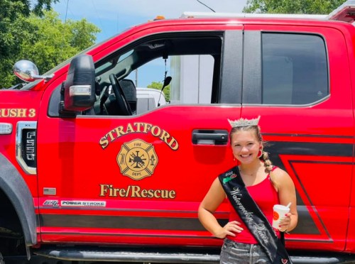 A smiling young woman in a crown and sash stands next to a red fire truck with "Stratford Fire/Rescue" written on it.