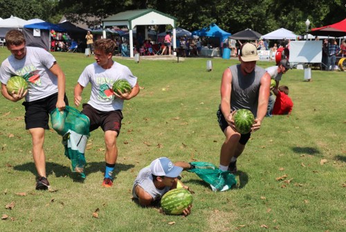 Four young men race while carrying watermelons, with spectators and tents in the background on a sunny day.