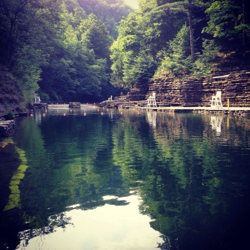 A serene lake surrounded by lush green trees and rocky cliffs, with a wooden path and lifeguard chairs in the background.