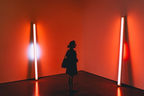 A person stands in a dimly lit room, illuminated by two vertical light fixtures in red and white hues.