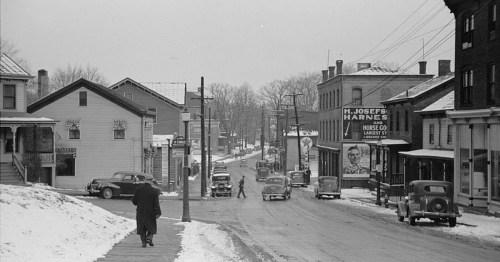 A snowy street scene from the past, featuring vintage cars, buildings, and a person walking along the sidewalk.