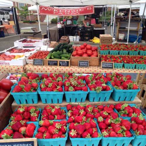 A vibrant farmers market display featuring baskets of strawberries, tomatoes, cucumbers, and other fresh produce.