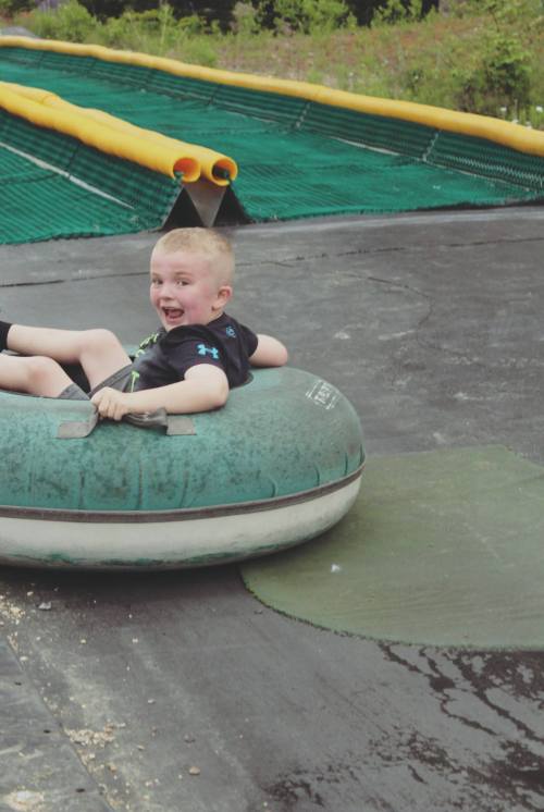 A smiling child sits in a green inner tube, ready to slide down a slope on a sunny day.