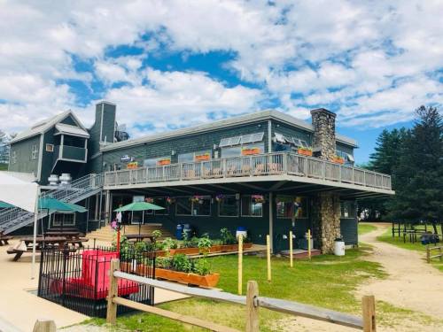 A green building with a deck, surrounded by trees and picnic tables, under a partly cloudy sky.