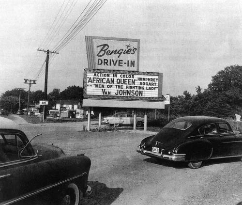 A vintage drive-in theater sign displaying movie titles, with classic cars parked nearby. Black and white photo.