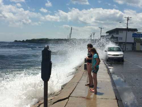 Two children stand by a waterfront, watching waves crash against the shore, with a car parked nearby.