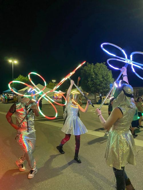Three performers in shiny outfits dance at night, wielding illuminated hula hoops and glowing props.