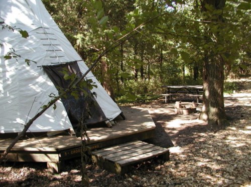 A white teepee beside a wooden deck in a wooded area, with a picnic table and fire pit nearby.