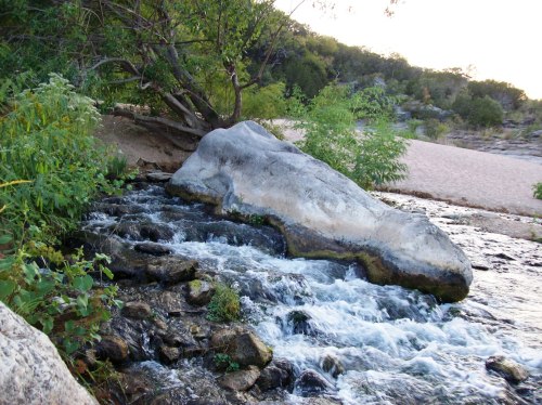 A serene stream flows over rocks, with lush greenery and a sandy bank in the background under a soft sunset light.