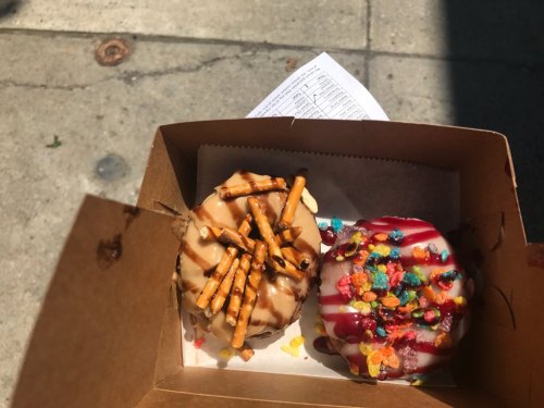 Two colorful donuts in a brown takeout box, one topped with pretzels and the other with fruity cereal and drizzle.