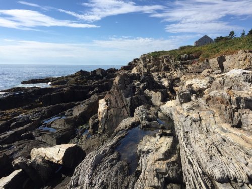 Rocky coastline with tide pools, under a blue sky with scattered clouds, and a house in the background.