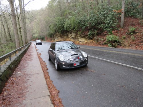 A small black car drives along a wet, winding road surrounded by trees and rocky terrain.
