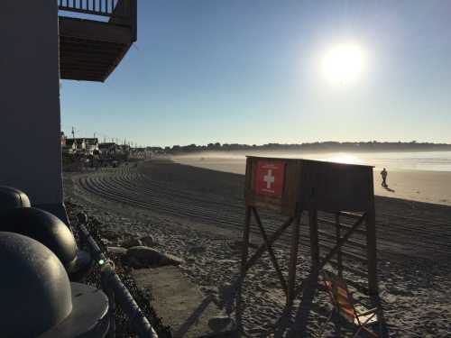 A beach scene at sunrise with a lifeguard station, smooth sand, and a distant figure walking along the shore.