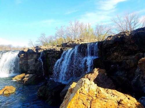 A scenic view of a waterfall cascading over rocky cliffs, surrounded by trees under a clear blue sky.