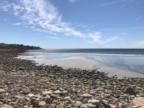 A rocky beach with calm waters under a clear blue sky and scattered clouds in the distance.