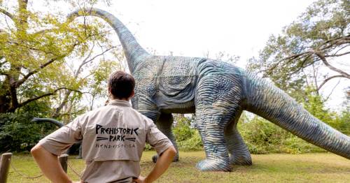 A person in a park uniform observes a large dinosaur statue in a lush outdoor setting.