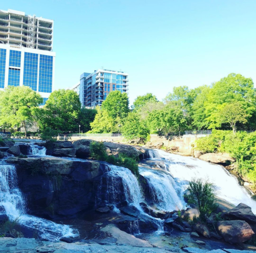 A scenic waterfall surrounded by greenery, with modern buildings in the background under a clear blue sky.