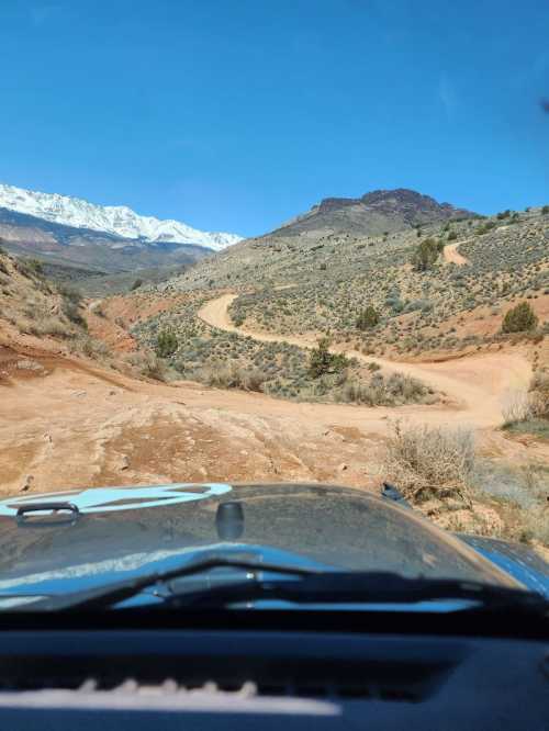 View from a vehicle on a winding dirt road surrounded by mountains and clear blue sky.