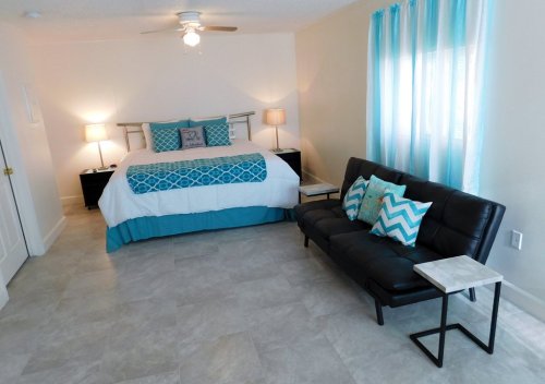Bright bedroom with a queen bed, teal accents, a black sofa, and large windows with sheer curtains.