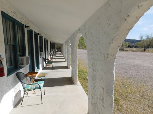 A row of motel rooms with white walls and blue trim, featuring chairs on a covered porch and a gravel parking area.