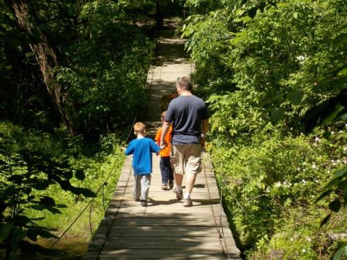 A man walks with two children on a wooden bridge surrounded by lush greenery.