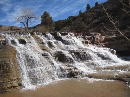 A cascading waterfall flows over rocky steps, surrounded by trees and a clear blue sky.