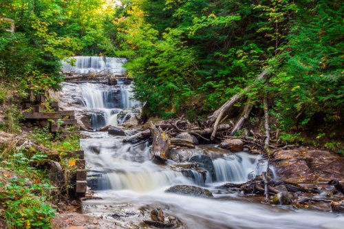 A serene waterfall cascades over rocks, surrounded by lush green trees and autumn foliage.