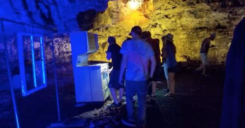 A group of people inside a cave, with a blue-lit kitchen setup and rocky walls in the background.