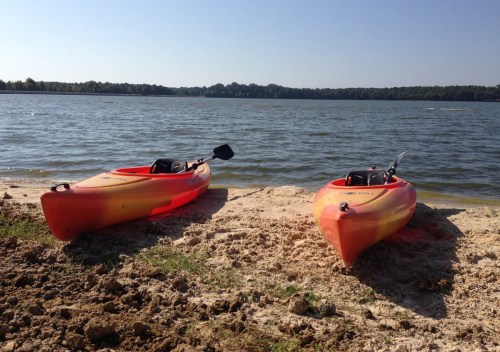 Two orange kayaks sit on a sandy shore by a calm lake under a clear blue sky.