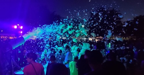 A lively night scene with a crowd enjoying a foam party, colorful lights illuminating the foam and people dancing.