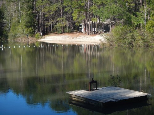 A serene lake scene with a wooden dock, surrounded by trees and a sandy shore in the background.
