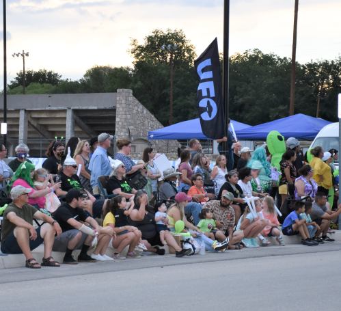 A diverse crowd sits along a street, some holding balloons, with a UFO flag and tents in the background.