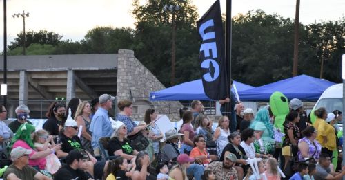 A crowd gathers at an outdoor event, with a UFO flag and tents in the background, and people of various ages enjoying the atmosphere.