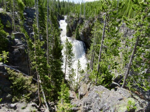 A waterfall cascades down rocky cliffs, surrounded by lush green trees in a scenic natural landscape.