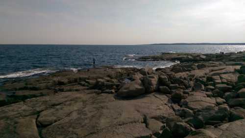 A person stands on rocky shorelines by the ocean, with gentle waves and a clear sky in the background.